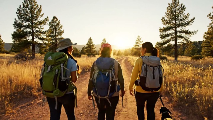 Women walking a dirt road into the low sun, a small dog ahead