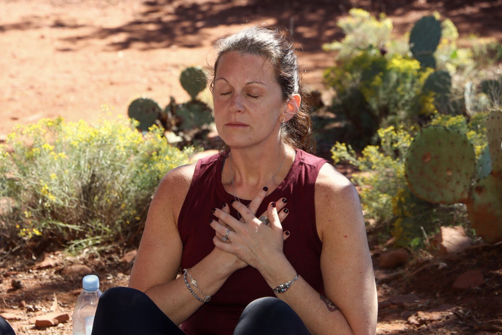 A woman seated in stillness against red rock, hand on heart, eyes closed in morning sun
